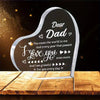 A clear acrylic heart plaque engraved with 'Dear Dad I love you even more' resting on a wooden surface.