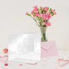 A clear acrylic gift box filled with pink carnations sits next to a pink envelope and scattered pink petals.
