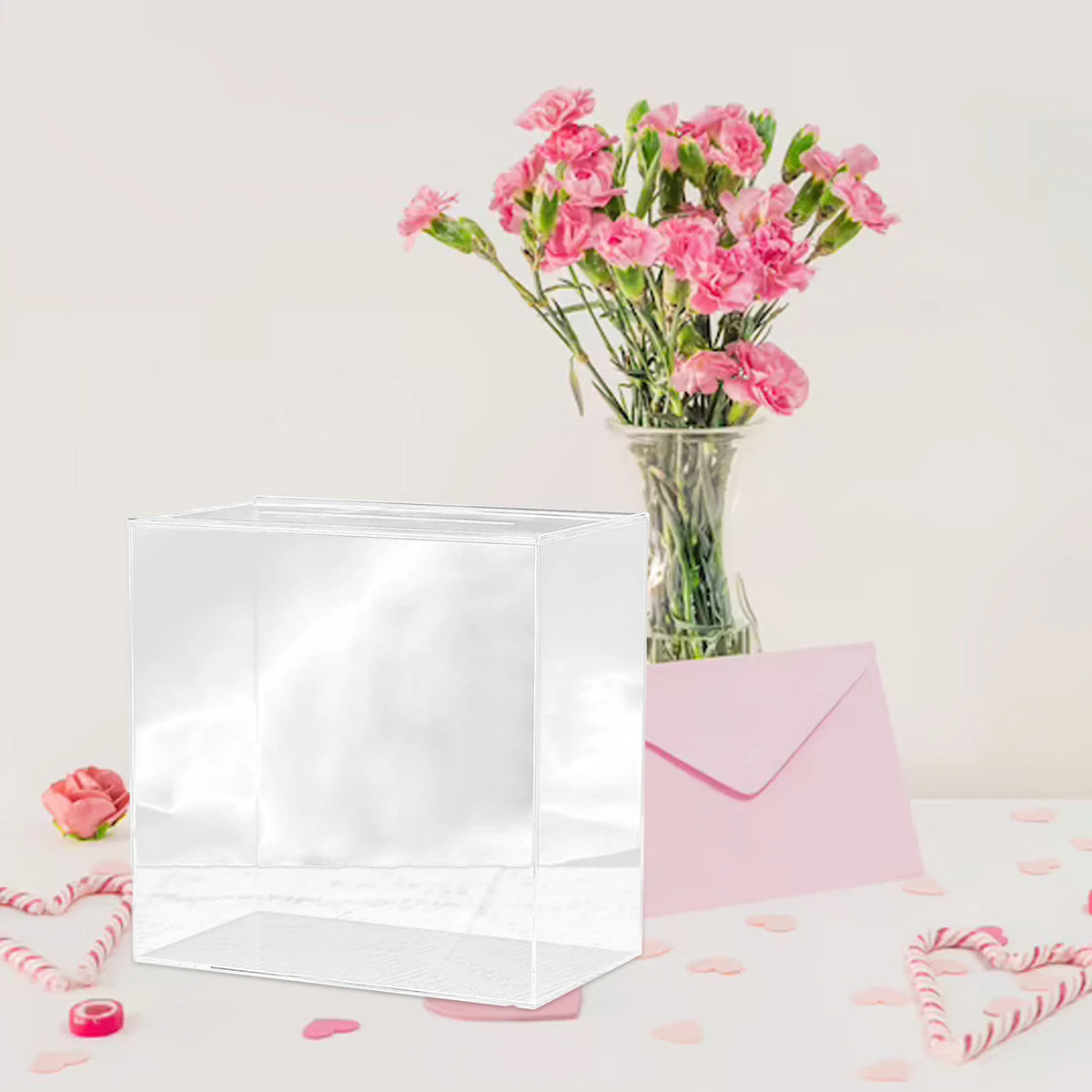 A clear acrylic gift box filled with pink carnations sits next to a pink envelope and scattered pink petals.