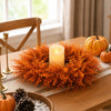 An orange artificial eucalyptus wreath with a white candle sits on a wooden table next to a pumpkin.