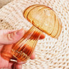A hand holds a translucent, mushroom-shaped glass vase with swirling amber and reddish tones against a woven background.