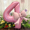A large pink foil balloon shaped like the number 4 with a matte bow, surrounded by green foliage and purple flowers.