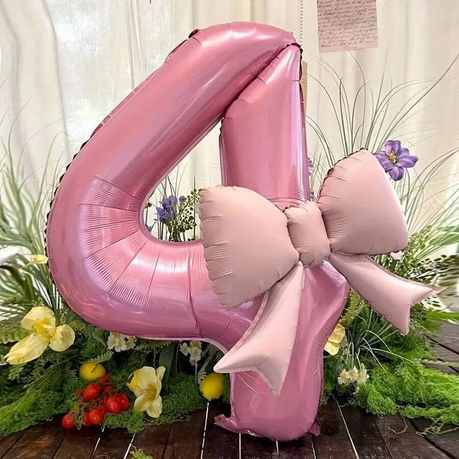 A large pink foil balloon shaped like the number 4 with a matte bow, surrounded by green foliage and purple flowers.