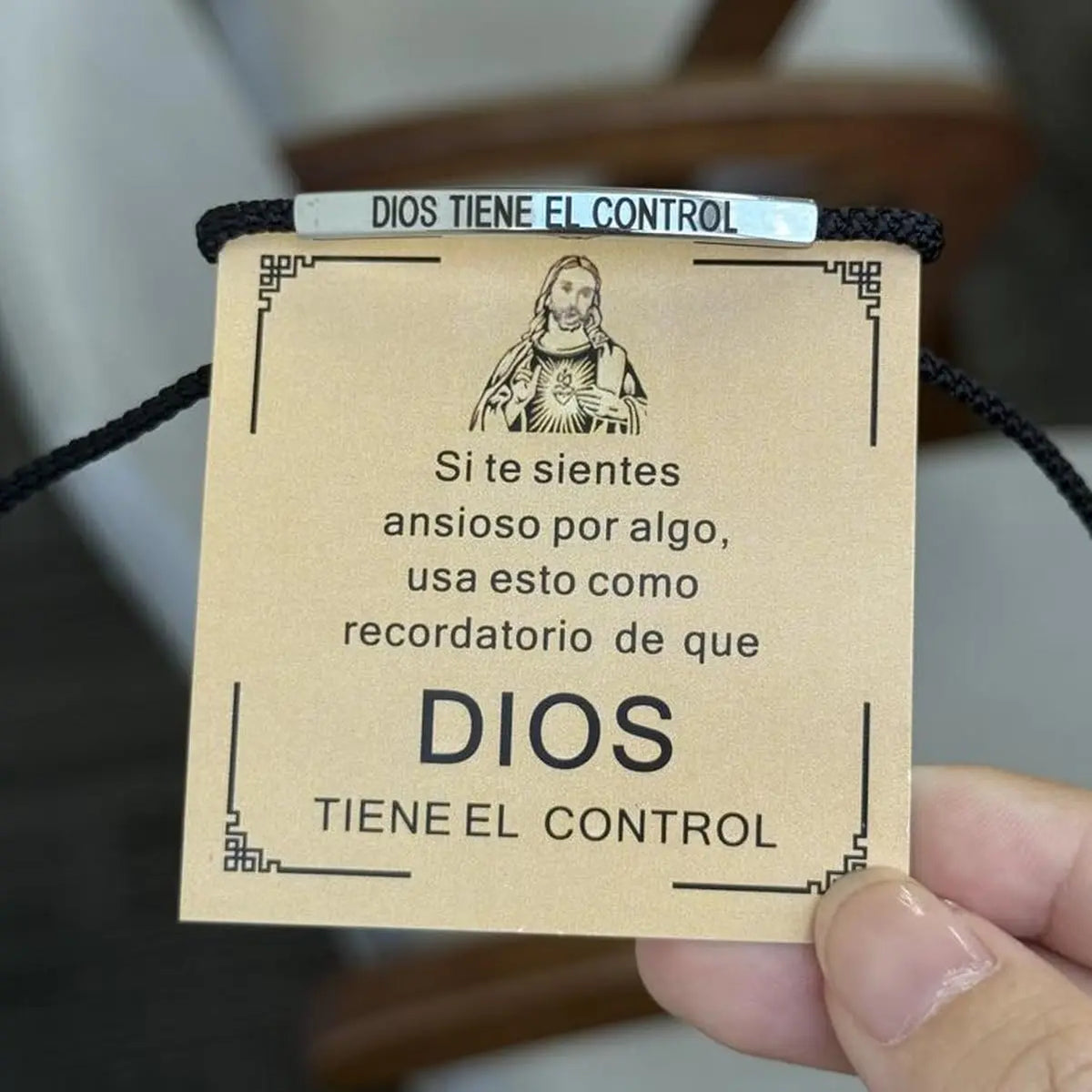 A close-up of a stainless steel bracelet with an engraved tag that reads Dios tiene el control and features a depiction of Je