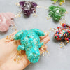A hand holds a bright turquoise crystal frog resin craft decoration surrounded by other colored frogs.