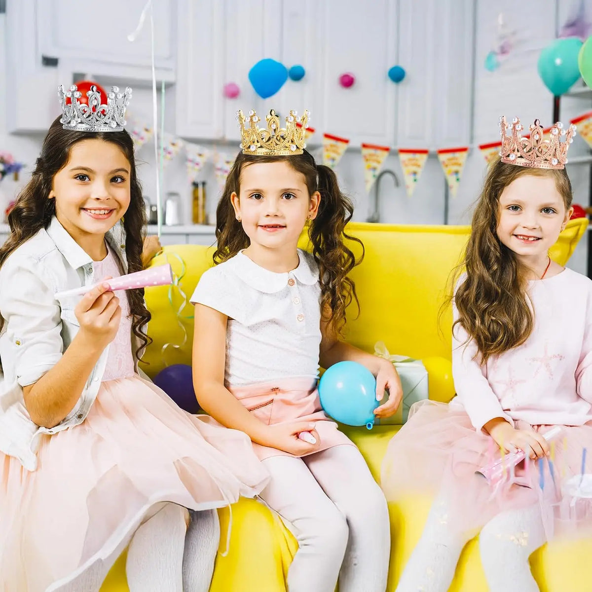 Three young girls wearing pearl crown cake toppers sitting on a yellow couch