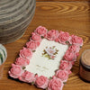 A pink rose resin picture frame with faux roses is displayed on a wooden surface.