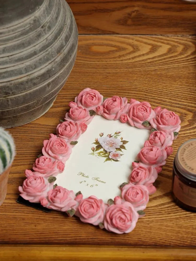 A pink rose resin picture frame with faux roses is displayed on a wooden surface.