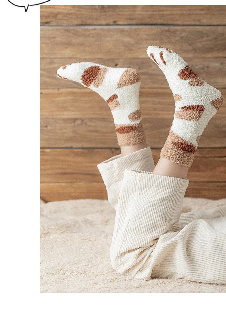 A pair of beige plush socks with brown cat paw prints are shown resting on a light-colored rug.