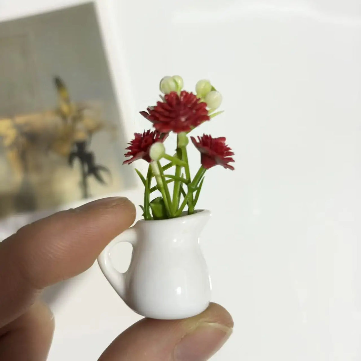 A hand holds a white ceramic vase with artificial red and white flowers, used as a fridge magnet.