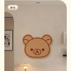 A round, brown cartoon bear felt wall decor panel hanging on a light beige wall above a table lamp.