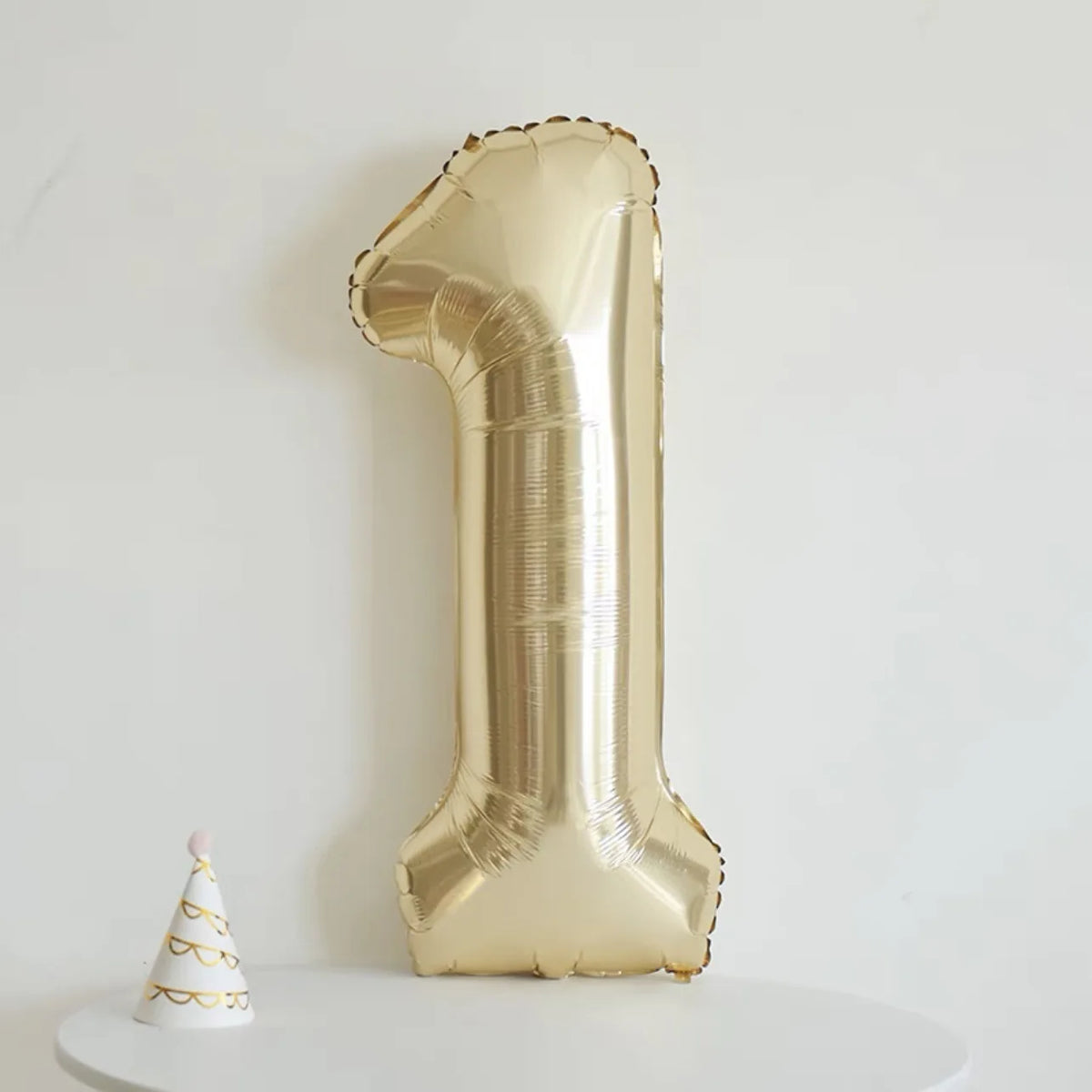 A large champagne gold foil number balloon shaped like the number one standing upright against a white wall.