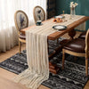 A wooden dining table set for a meal with a sheer beige cheesecloth table runner and dark patterned rugs.