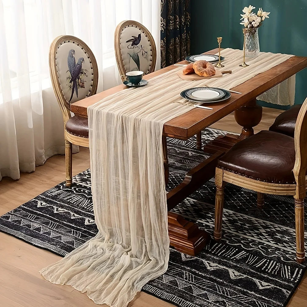 A wooden dining table set for a meal with a sheer beige cheesecloth table runner and dark patterned rugs.