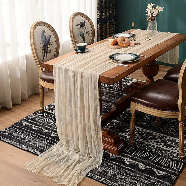 A wooden dining table set for a meal with a sheer beige cheesecloth table runner and dark patterned rugs.