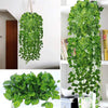 A lush green artificial ivy vine garland hanging on a white wall, shown alongside a potted plant.