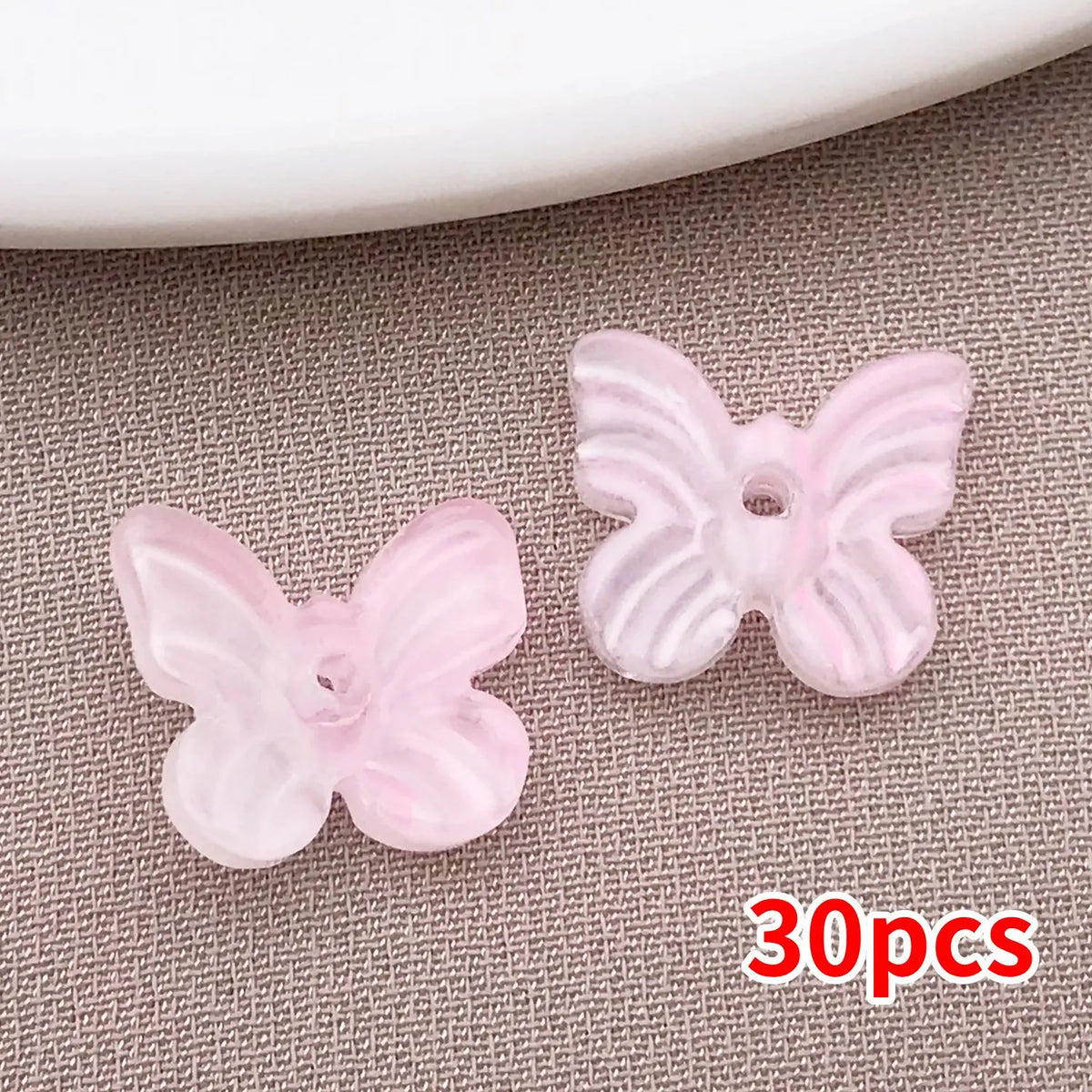 Un juego de 30 cuentas de mariposa de vidrio rosa sobre una superficie gris texturizada.