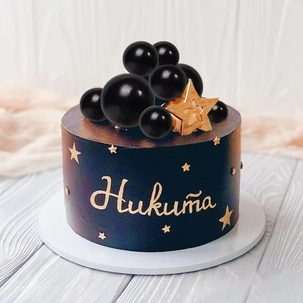 A dark blue cake decorated with gold stars and black spherical toppers, featuring the name Hukuma.