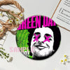 A circular button pin featuring a stylized face with pink X marks on his eyes against a green background.