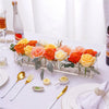 Clear acrylic rectangular vase filled with an arrangement of orange and yellow roses on a white tablecloth.
