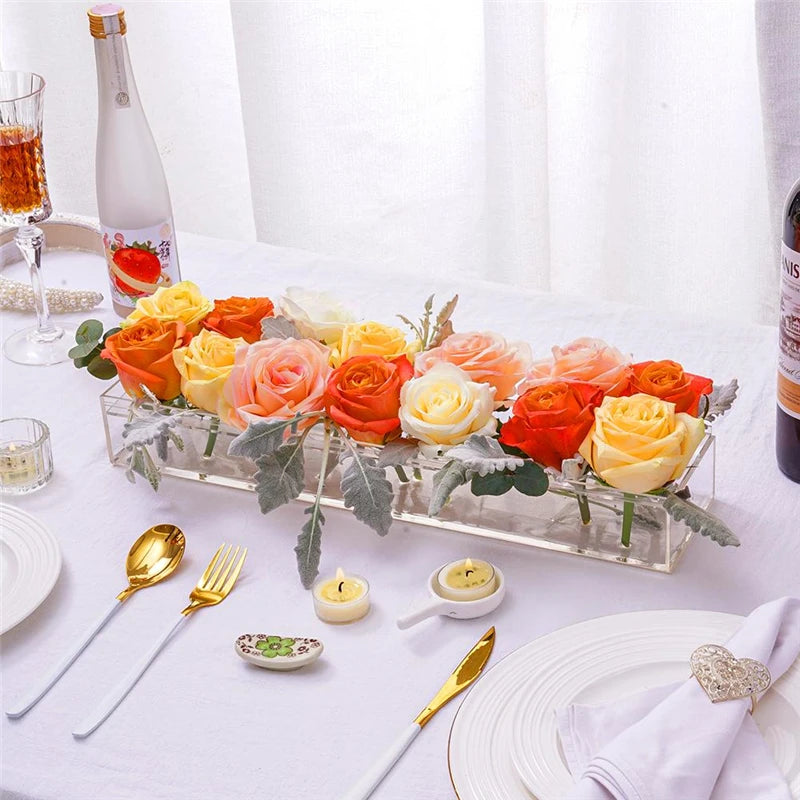 Clear acrylic rectangular vase filled with an arrangement of orange and yellow roses on a white tablecloth.