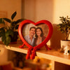 A red heart-shaped frame with a reflective surface holds a photo of a smiling couple.