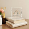 Acrylic nativity scene Christmas ornament displayed on a wooden surface next to a stack of books.