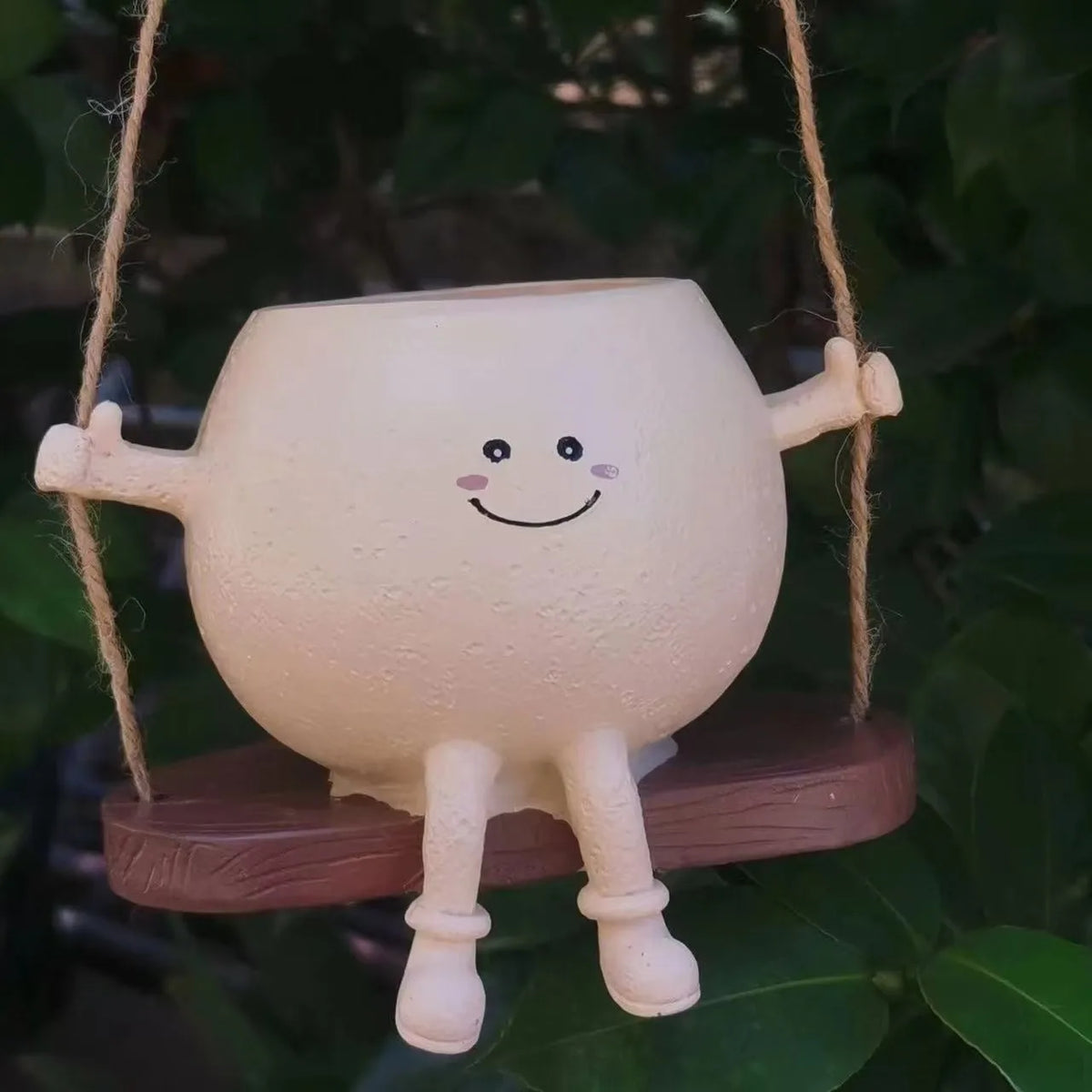 A white resin ornament shaped like a smiling figure is hanging from twine against a dark green background.