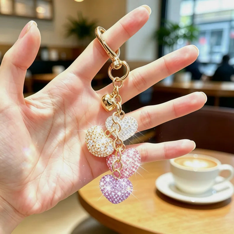 A hand holds a gold-colored crystal heart rhinestone keychain and bag charm.