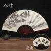 A black and white traditional folding fan with a landscape painting design is displayed on a wooden surface.