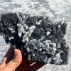 A hand holds a cluster of natural zinc crystals with iridescent white and gray tones.