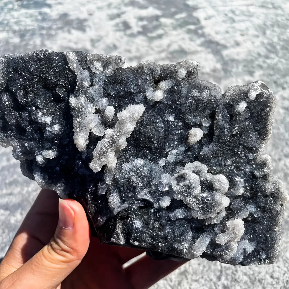 A hand holds a cluster of natural zinc crystals with iridescent white and gray tones.