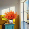 A potted artificial fall eucalyptus and flower stems display bright orange-red foliage on a wooden table near a window.