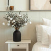 A large matte black decorative vase filled with silvery-green foliage and white fluffy accents sits on a light wood shelf.