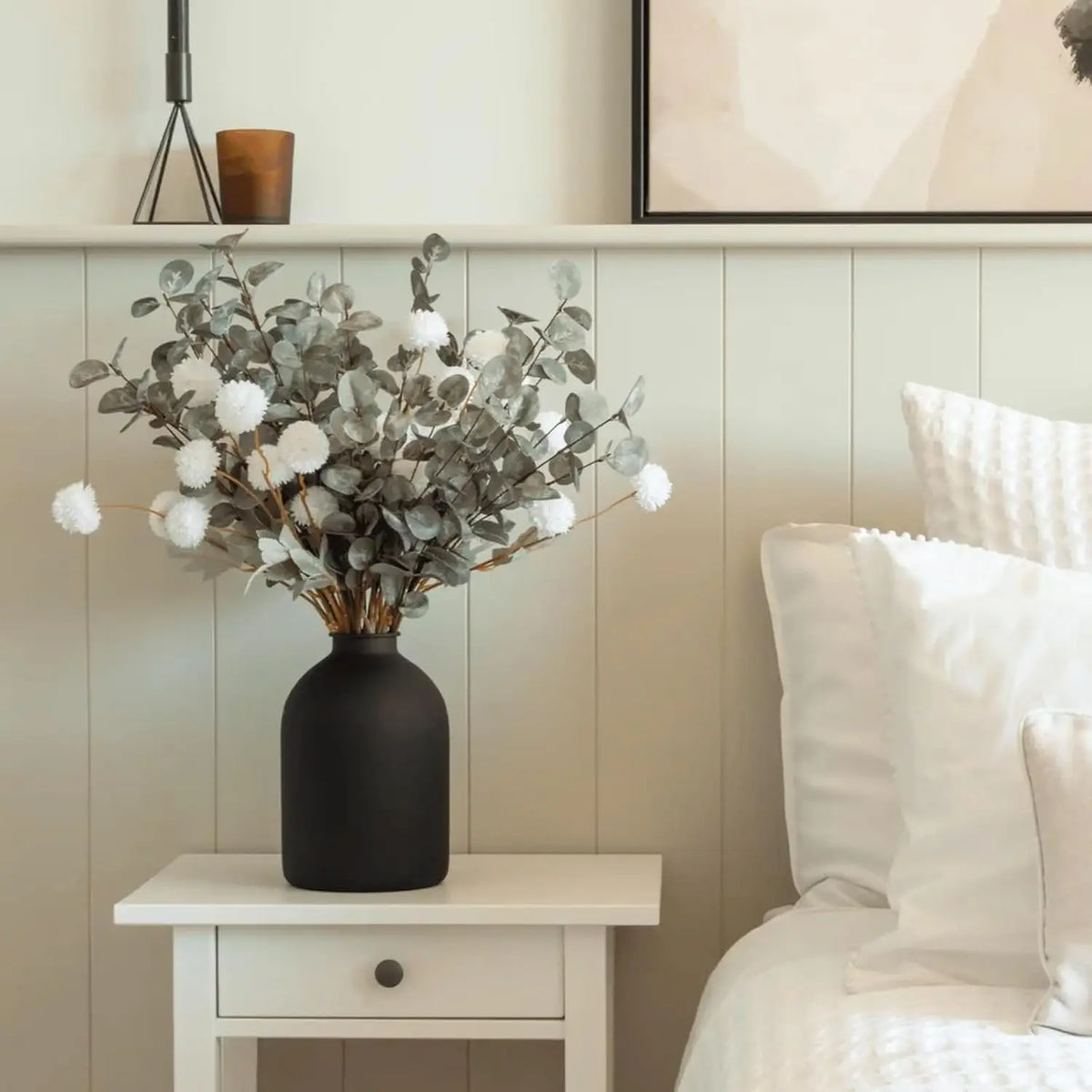 A large matte black decorative vase filled with silvery-green foliage and white fluffy accents sits on a light wood shelf.