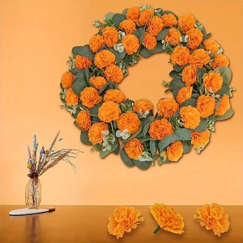 A wreath of orange carnation and marigold flower heads with greenery displayed against an orange wall.