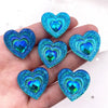 A hand holds a set of twelve heart-shaped blue iridescent peacock resin rhinestone flatbacks.