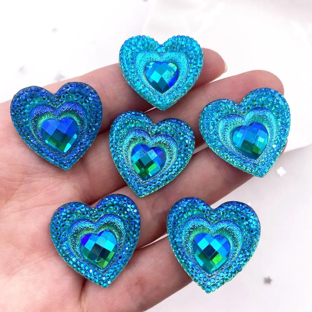 A hand holds a set of twelve heart-shaped blue iridescent peacock resin rhinestone flatbacks.