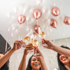 Hands lift champagne flutes filled with golden liquid beneath a balloon garland