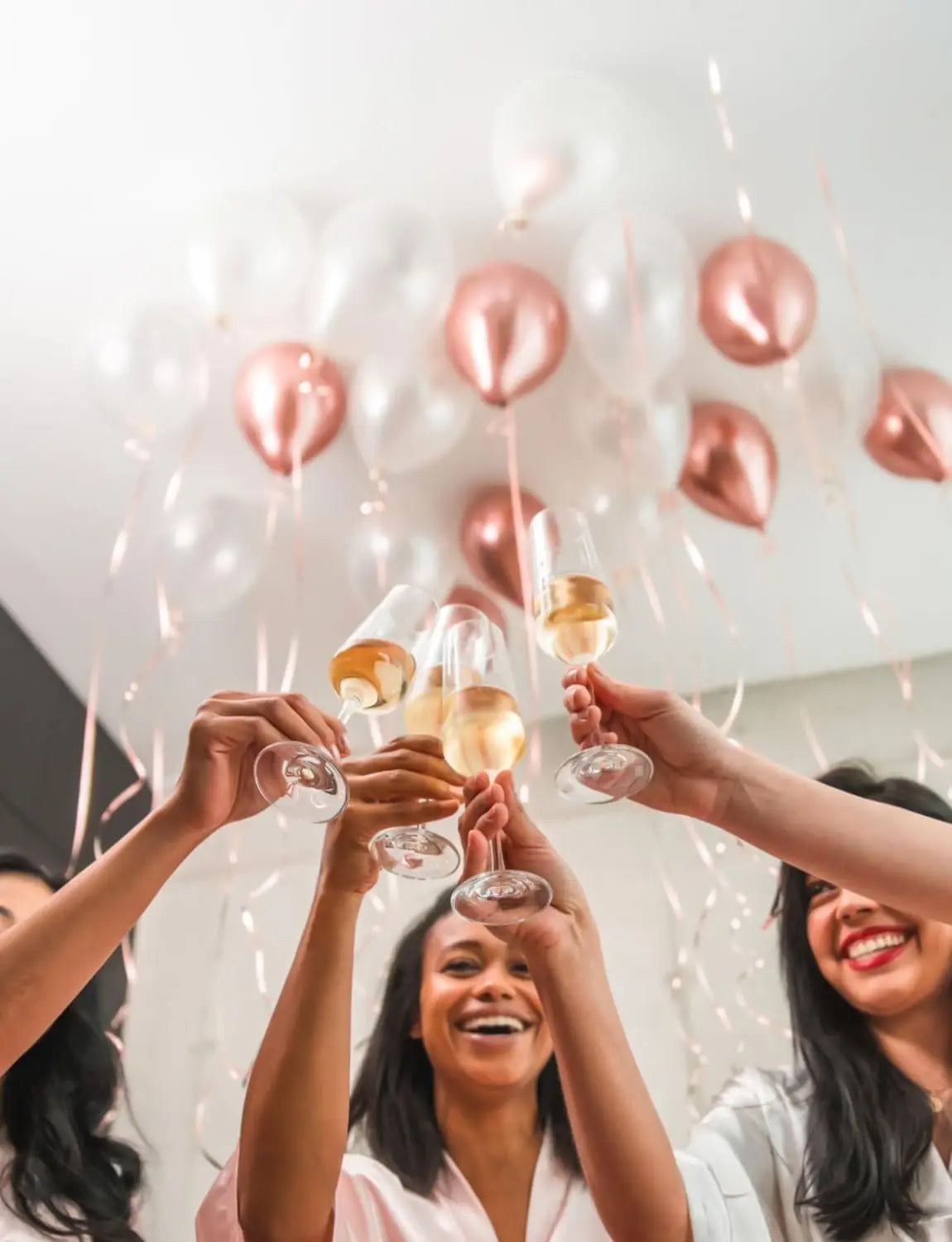 Hands lift champagne flutes filled with golden liquid beneath a balloon garland