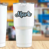 A white tumbler with a black script name Mark printed on it, set against a blurred background.