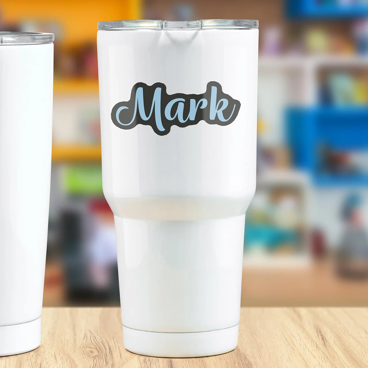 A white tumbler with a black script name Mark printed on it, set against a blurred background.