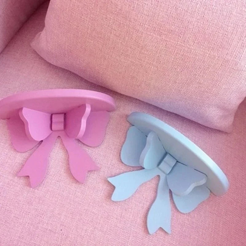 Two pink and blue decorative bow-shaped wooden wall shelves resting on a pink fabric surface.