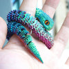 A hand holds a vibrant, textured 3D printed articulated snake fidget toy with iridescent green and purple scales.