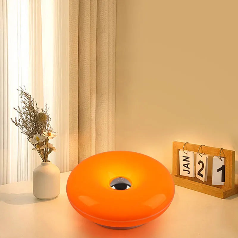 Orangene, donutförmige, wiederaufladbare Wandlampe auf weißer Fläche neben durchscheinenden Vorhängen.