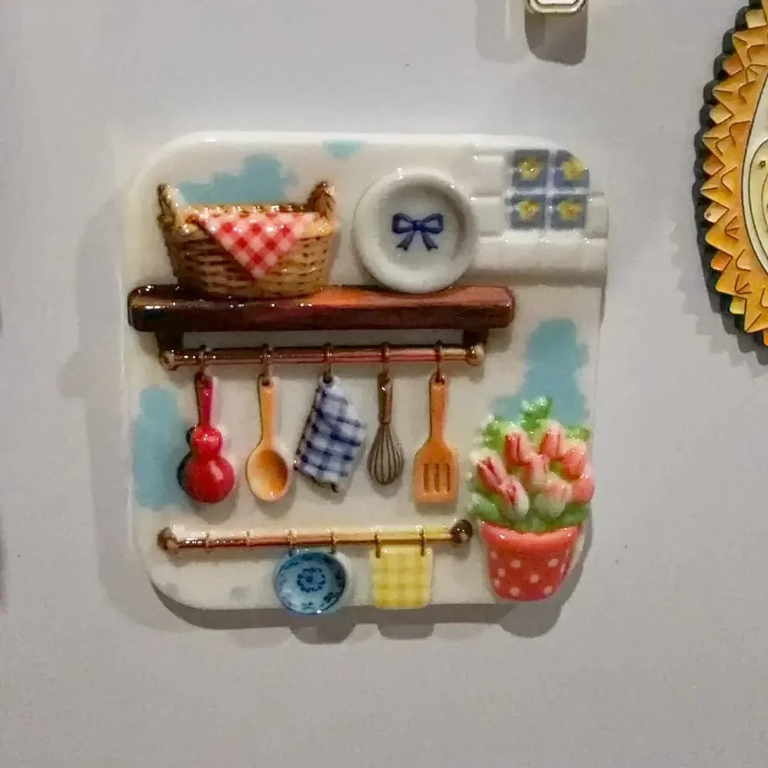 A white resin fridge magnet featuring embossed kitchen items like a woven basket, a small plate, and various utensils on a wo