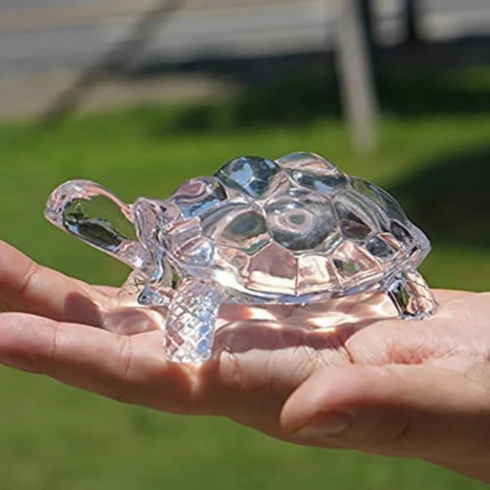 A clear crystal turtle desk ornament is held in a person's hand against a blurred green outdoor background.