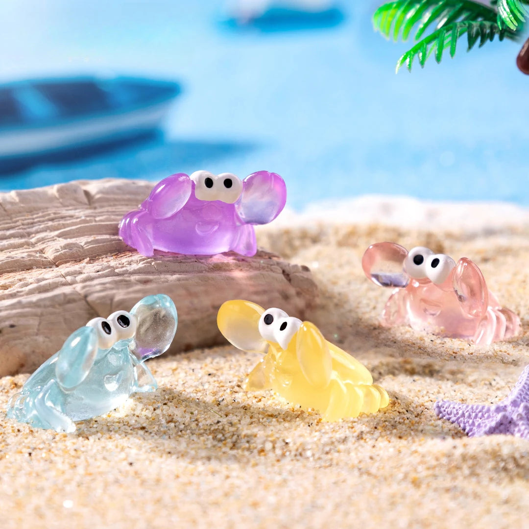 A set of luminous resin mini crab decor in purple, pink, and yellow resting on light sand.