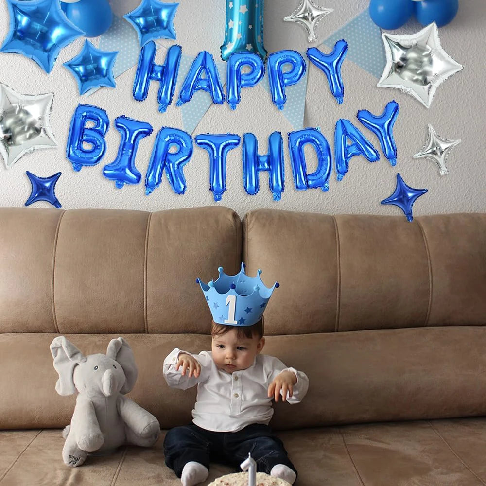 A baby boy wearing a blue crown sits in front of a wall decorated with blue and silver foil balloons for a birthday celebrati