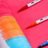A set of dual-ended watercolor art markers showing various bright colors like orange, blue, and pink on a pink background.